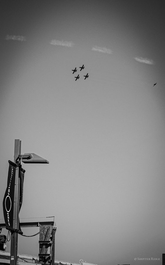 Four planes in formation across a vast grey sky above a street lamp, black and white