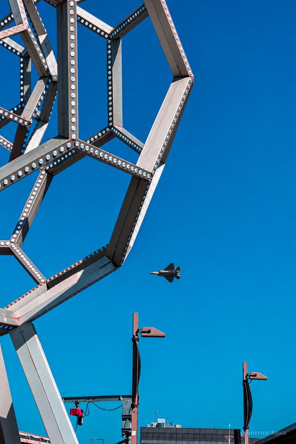 Polyhedral lattice sculpture against a deep blue sky with a jet passing